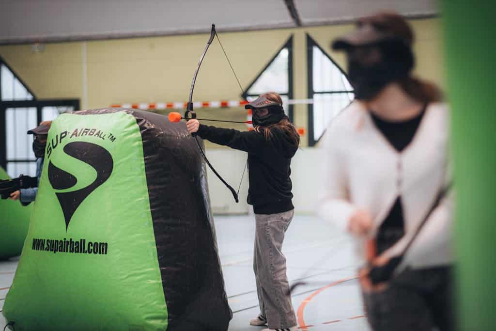 Players participating in an archery tag game indoors, aiming with bows and arrows.