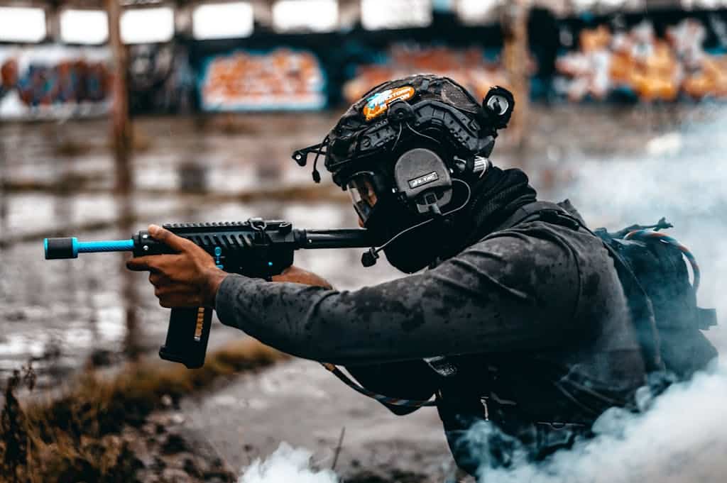 An airsoft player equipped with a helmet and tactical gear aiming a replica gun amid smoke.
